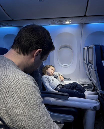 Parent and child on a long-haul flight, the child resting on a Travel Nest Kids Bed by the window
